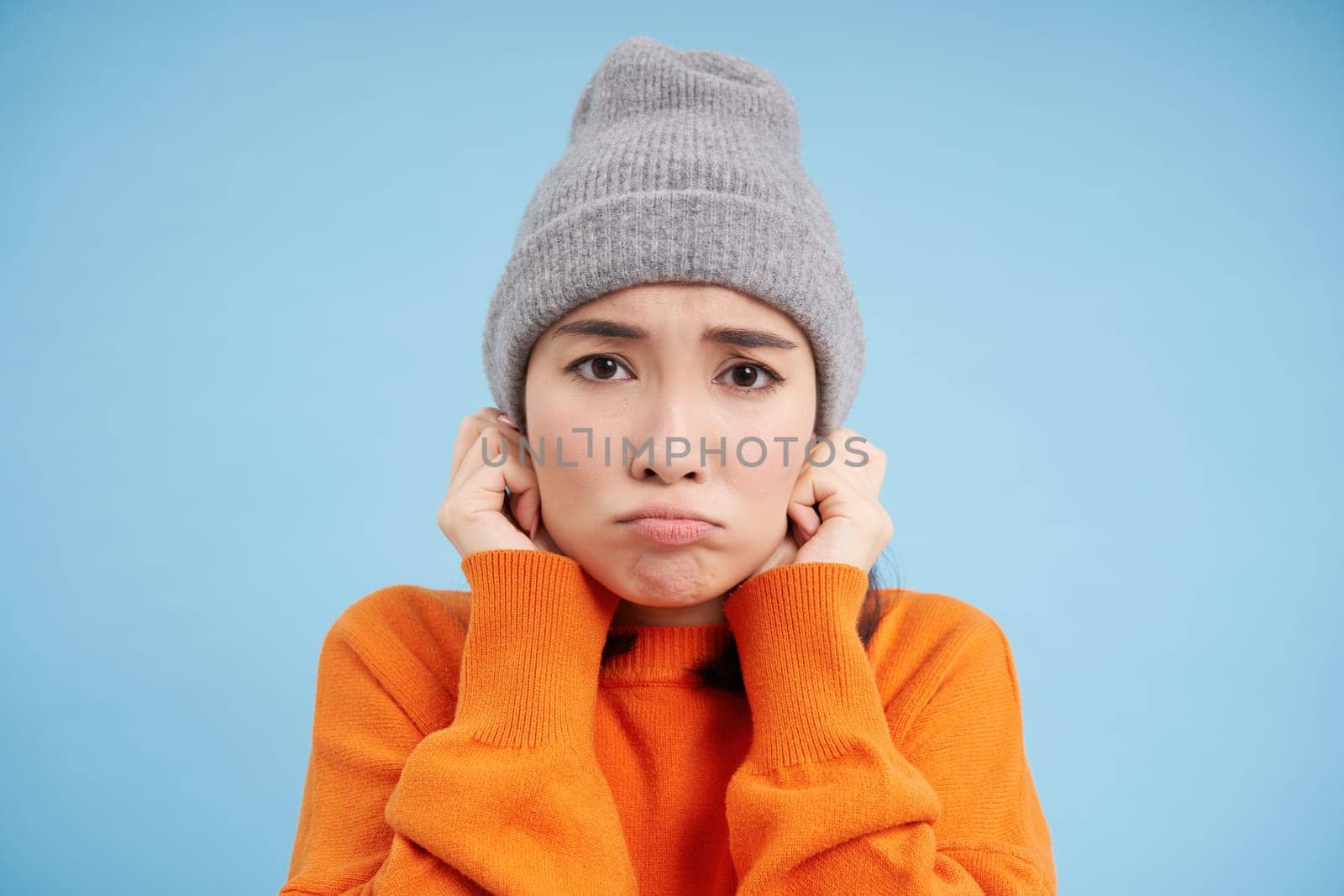 Close up portrait of korean girl feels cold, shakes and freezes, puts on beanie and sulks with sad face, stands over blue background.