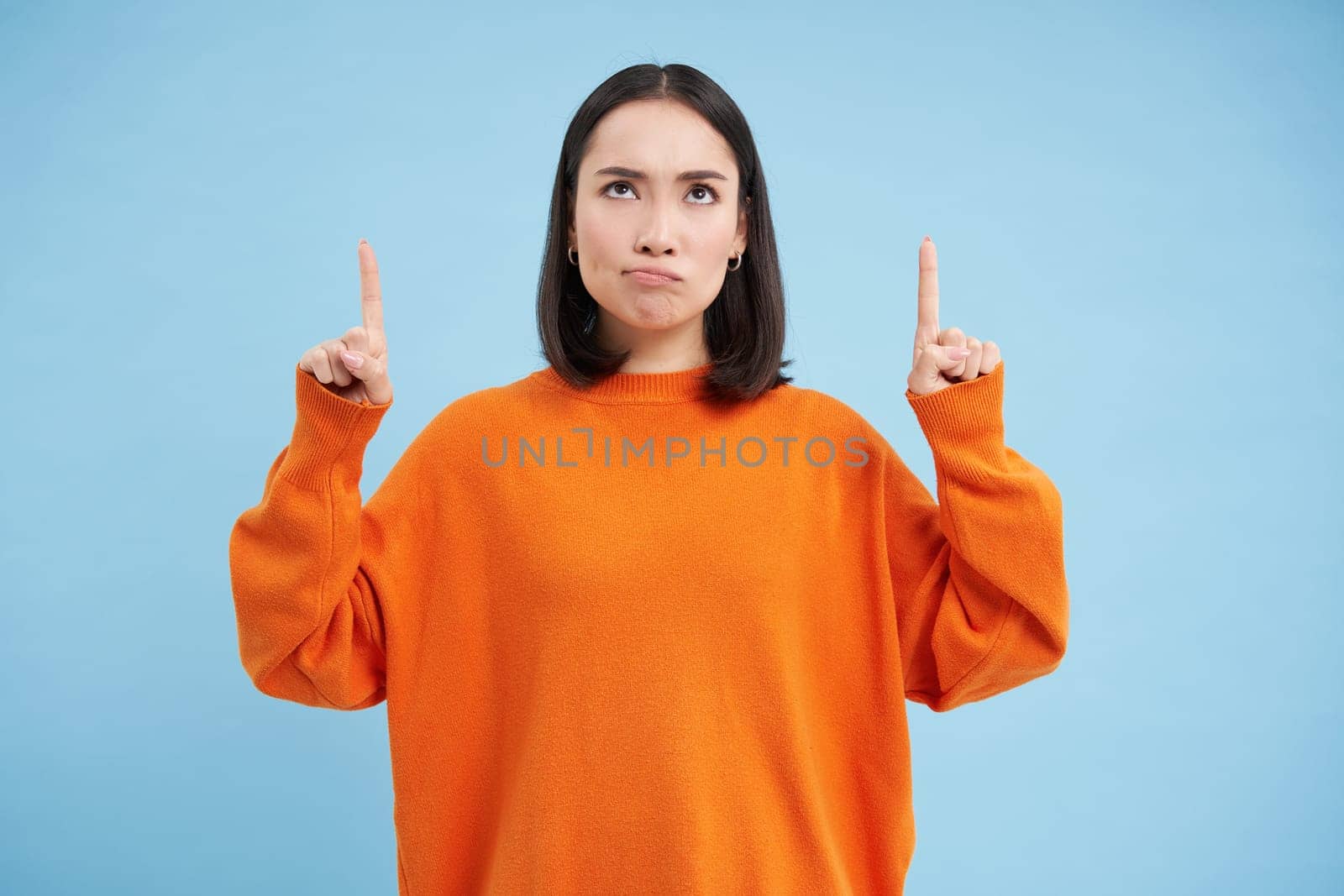 Perplexed asian girl, looks and points up with frustrated, thinking face expression, shows advertisement and reads it with doubt, stands over blue background.