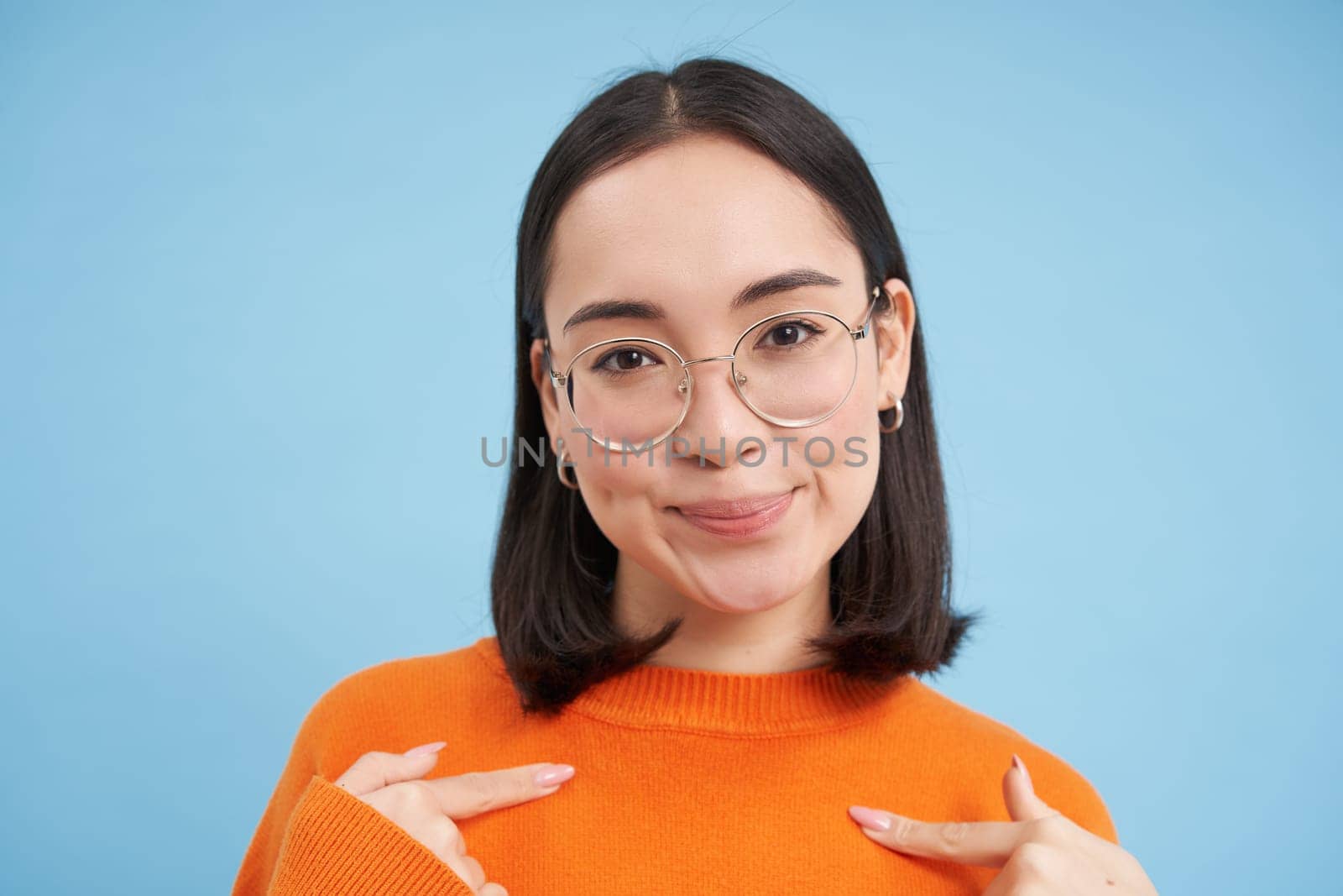 Portrait of beautiful girl in stylish glasses, pointing at herself with confidence, being self-assured, standing over blue background.
