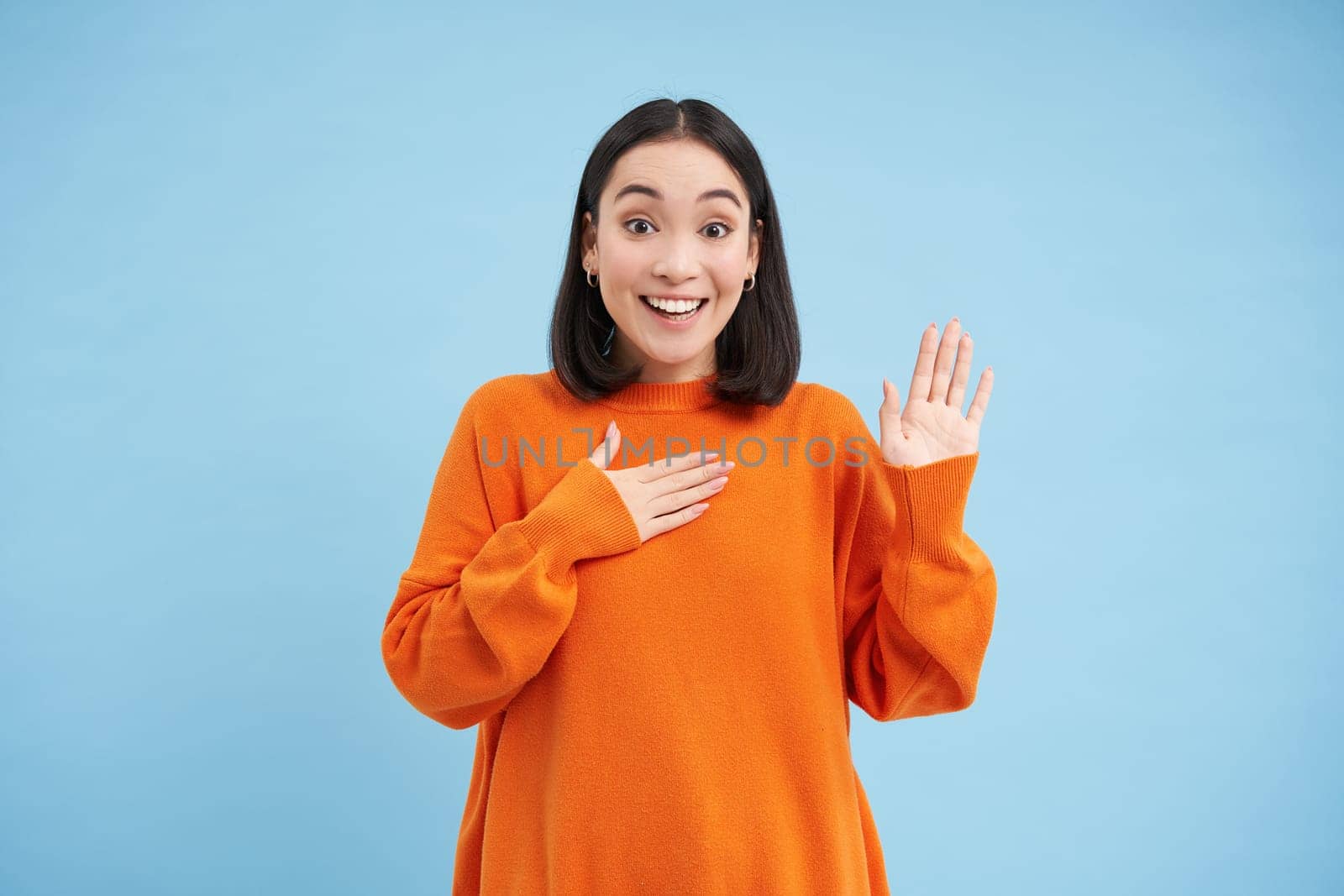 Smiling asian woman press arm to heart and raises one hand, introduces herself, stands over blue background.