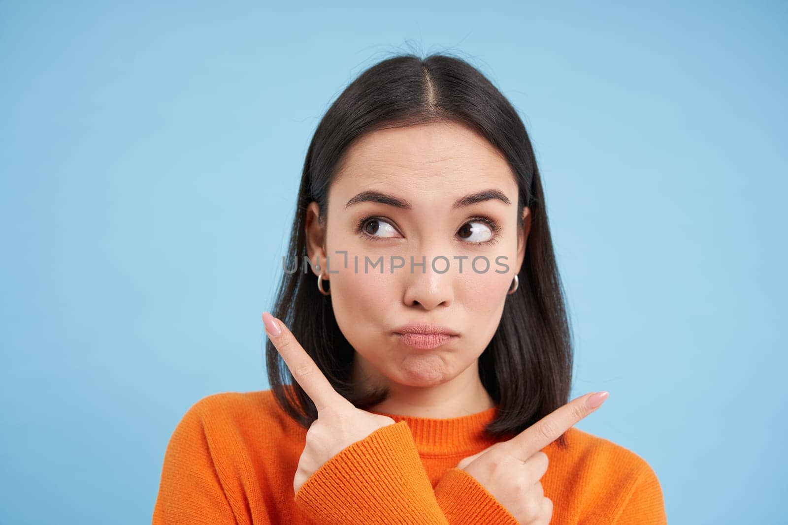 Close up of puzzled asian girl, points left and right, shows two choices, makes decision, stands over blue background by Benzoix
