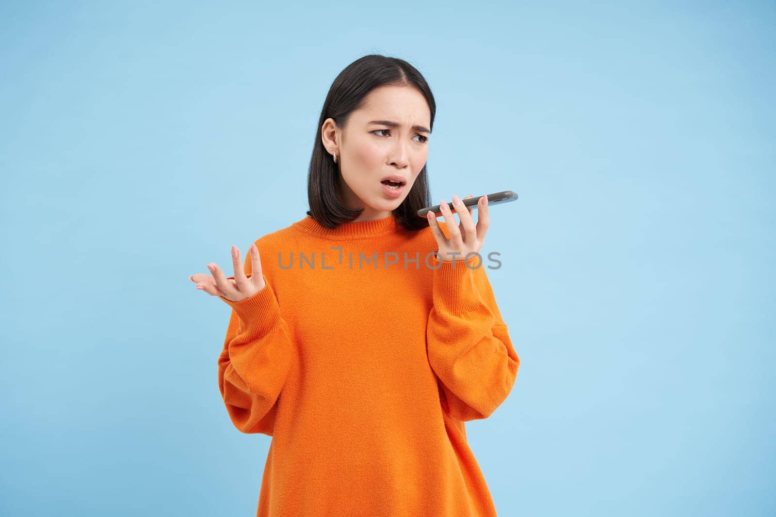 Portrait of upset young woman records voice mail or message, complains with disappointed, sad face, standing over blue background and holding mobile phone by Benzoix