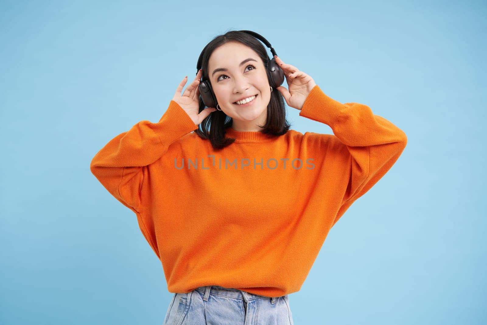 Happy Chinese woman in headphones, listens music, enjoys favourite song in her playlist, stands over blue background. Copy space