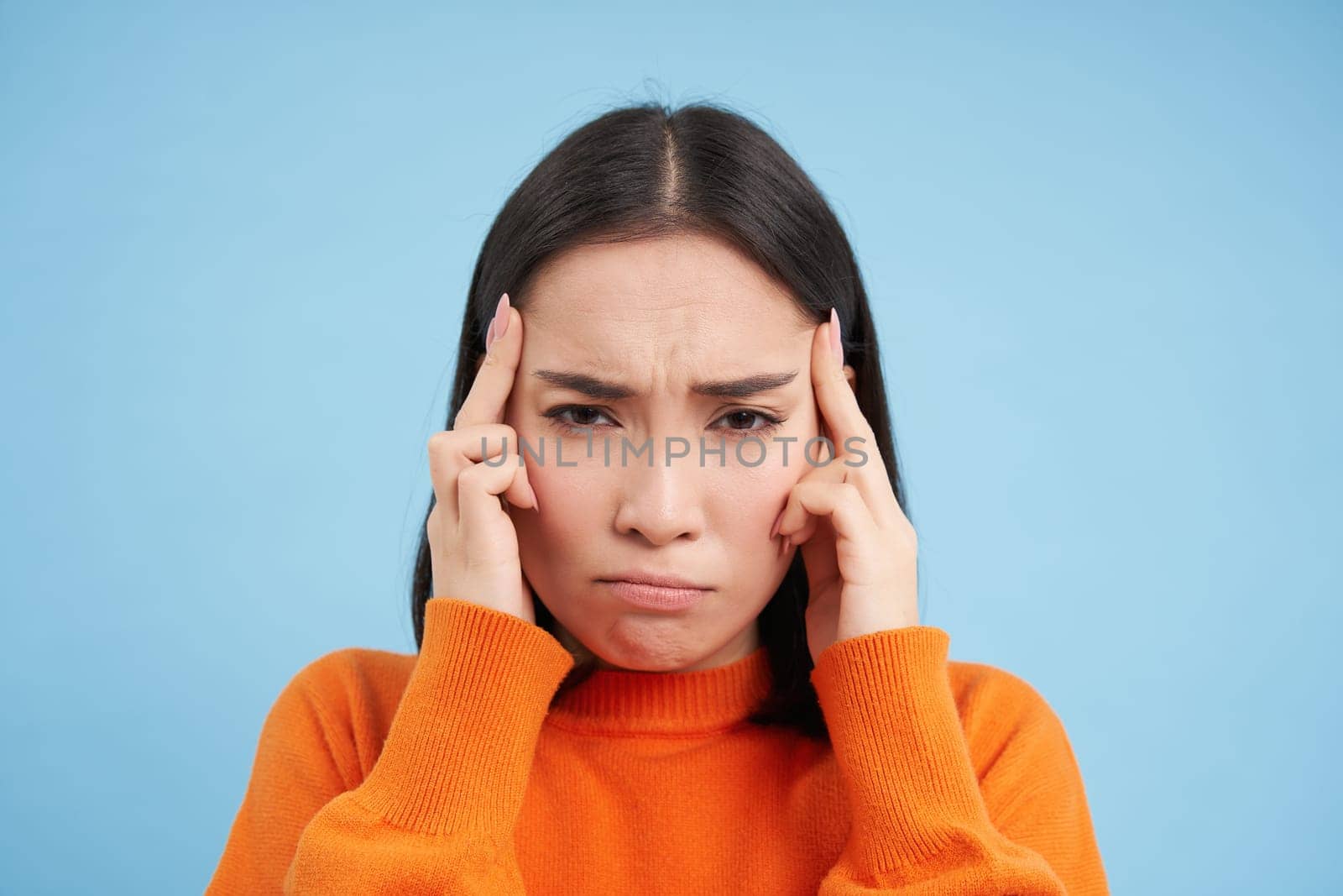 Close up portrait of asian woman with migraine, touches her head and massaging temples, has headache, stands over blue background.