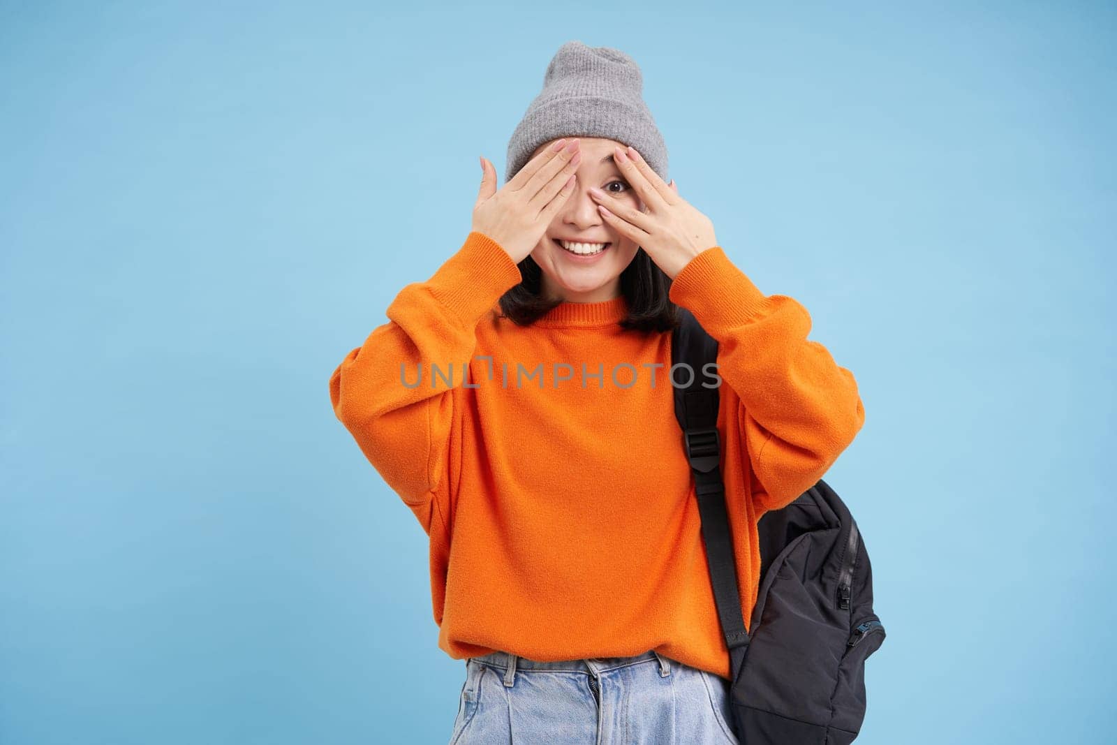 Enthusiastic girl, student closes her eyes, peeks through fingers and waits for surprise, stands over blue background.