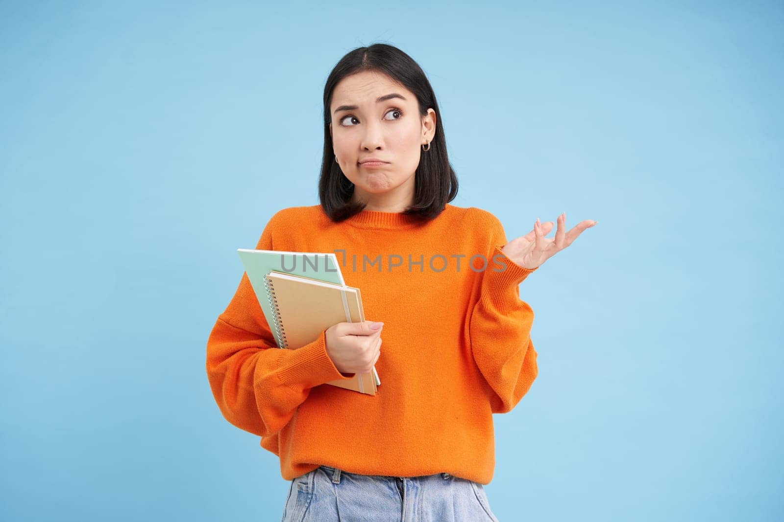 Confused asian woman, student with puzzled face shrugs shoulders, holds notebook, stands over blue background by Benzoix