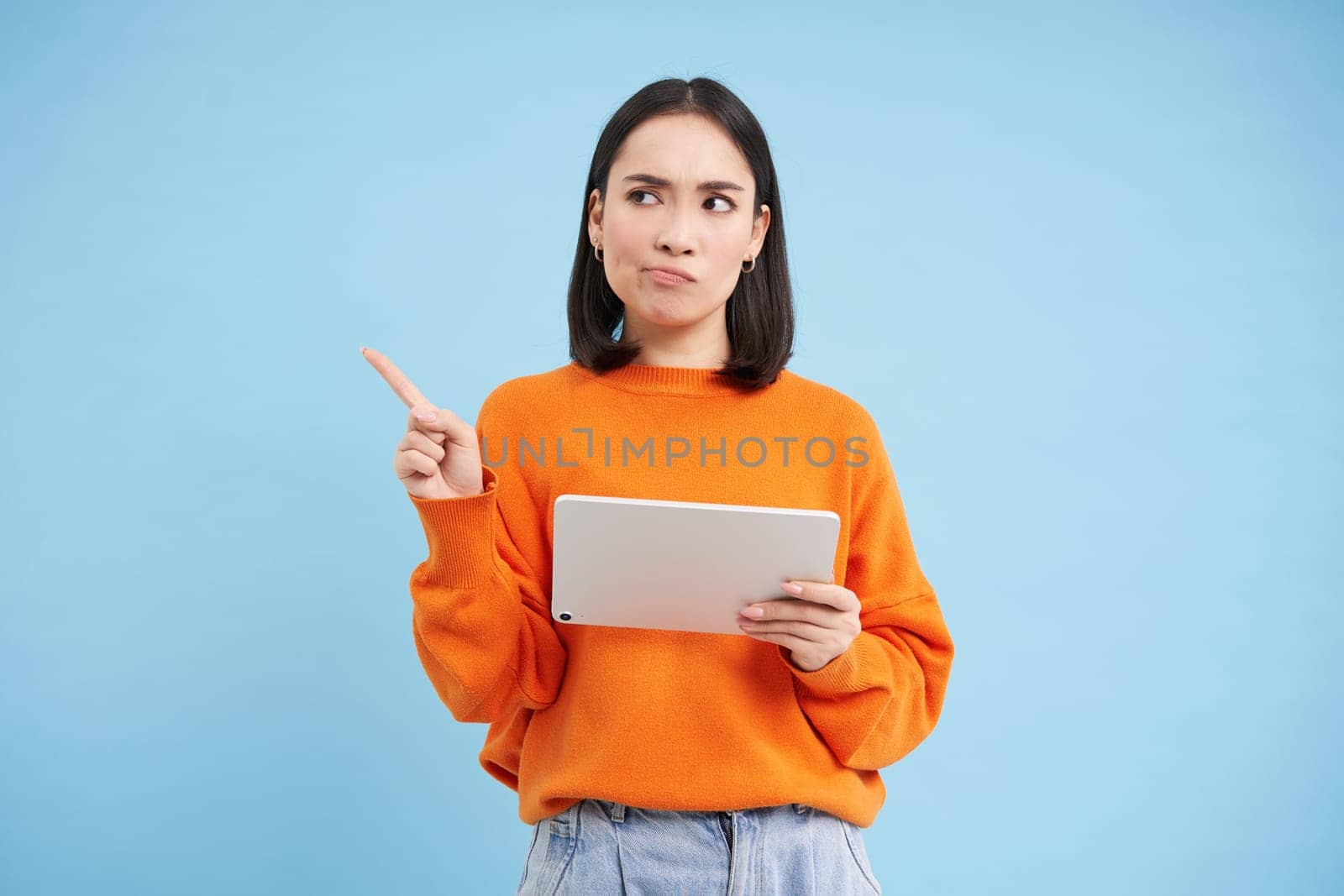 Portrait of korean woman with digital tablet, pointing at upper left corner with confused, frowning face, standing over blue background by Benzoix