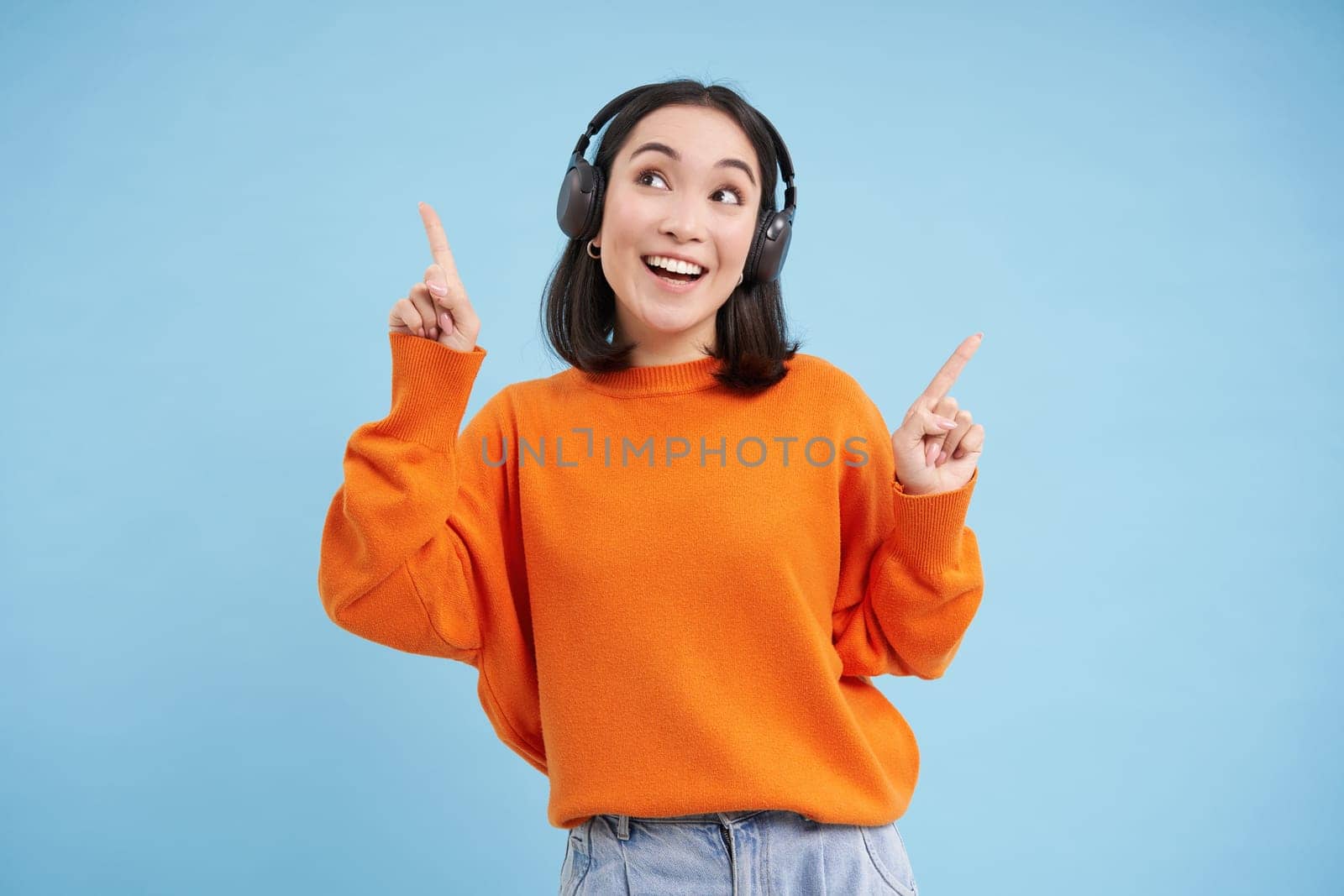 Happy Chinese woman in headphones, listens music, enjoys favourite song in her playlist, stands over blue background by Benzoix