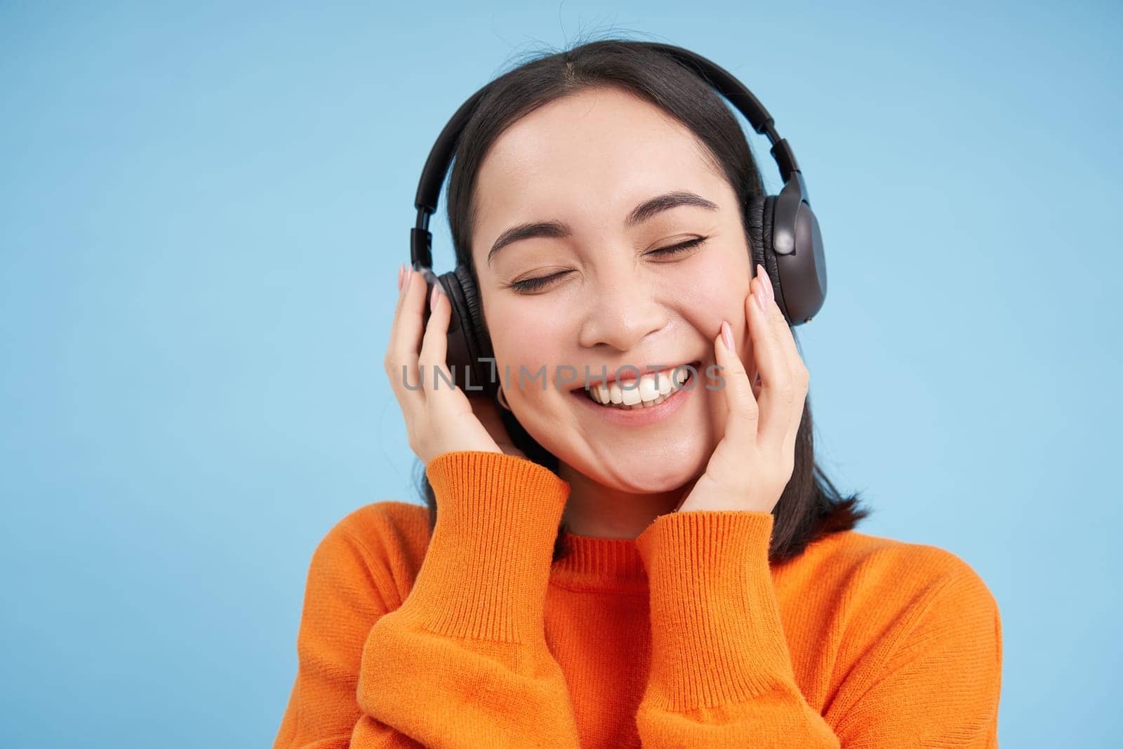 Close up of beautiful asian woman in headphones, smiles and listens music, enjoys podcast, stands over blue background by Benzoix