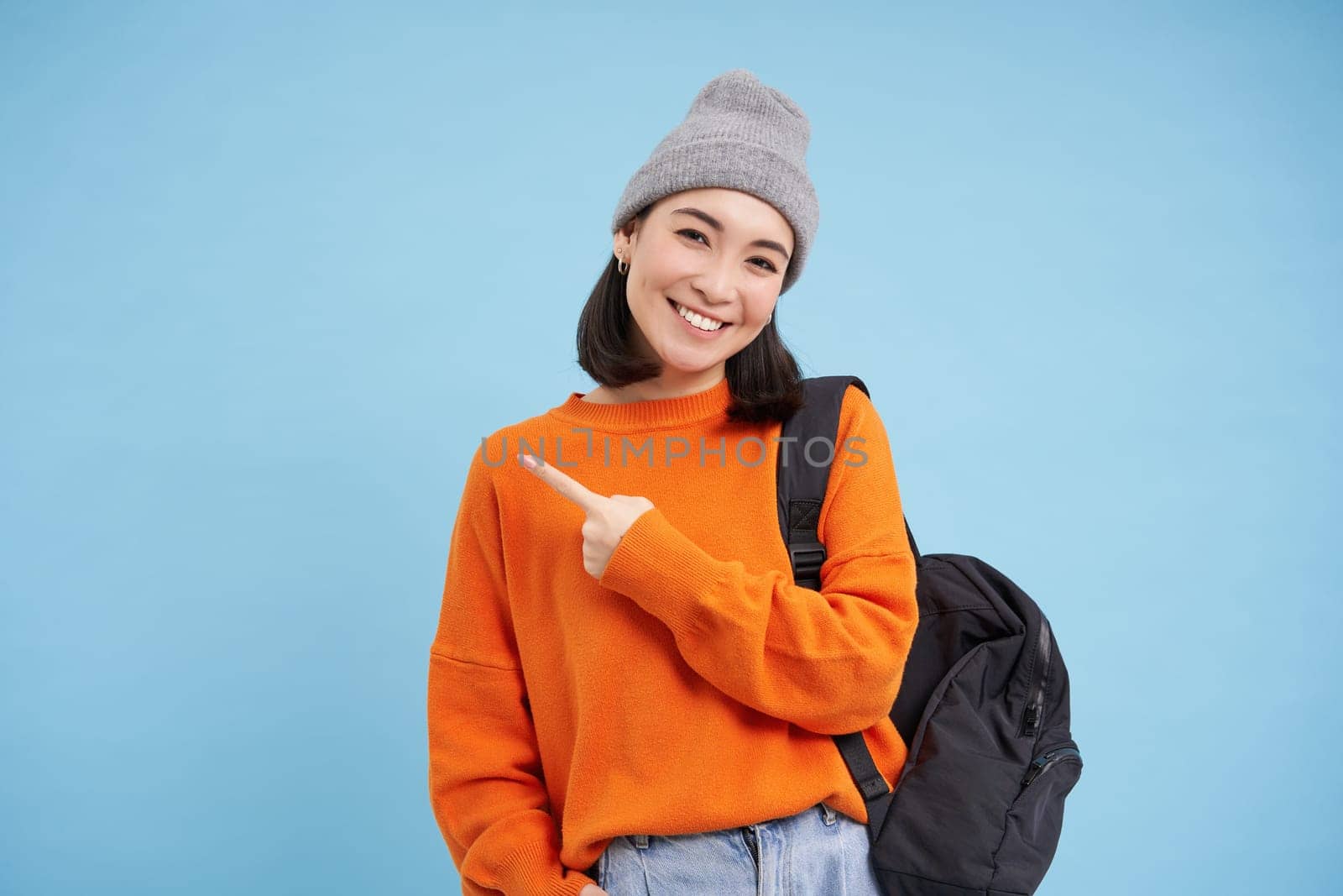Enthusiastic asian woman, points finger left at banner, shows promo offer, smiles and looks happy, stands in hat with backpack, blue background by Benzoix