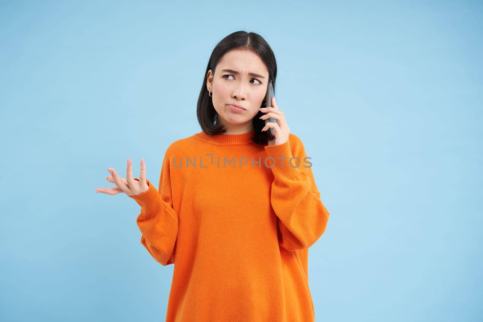 Sad and confused asia girl shrugs, talks on cellphone, answers telephone call with puzzled face, stands over blue background by Benzoix