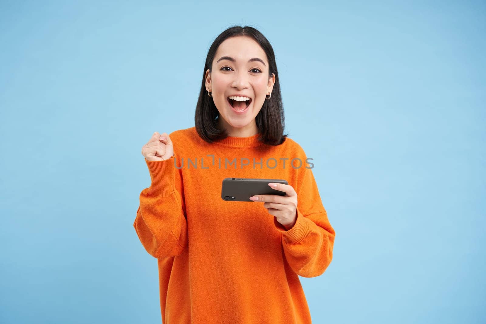 Excited Chinese girl wins on mobile video game, triumphing and laughing from joy, achieve goal on smartphone application, blue background by Benzoix