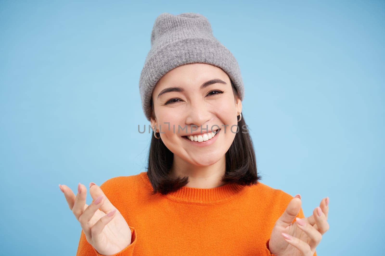 Close up portrait of happy smiling asian woman shows open hands, pointing, looking friendly, standing over blue background by Benzoix