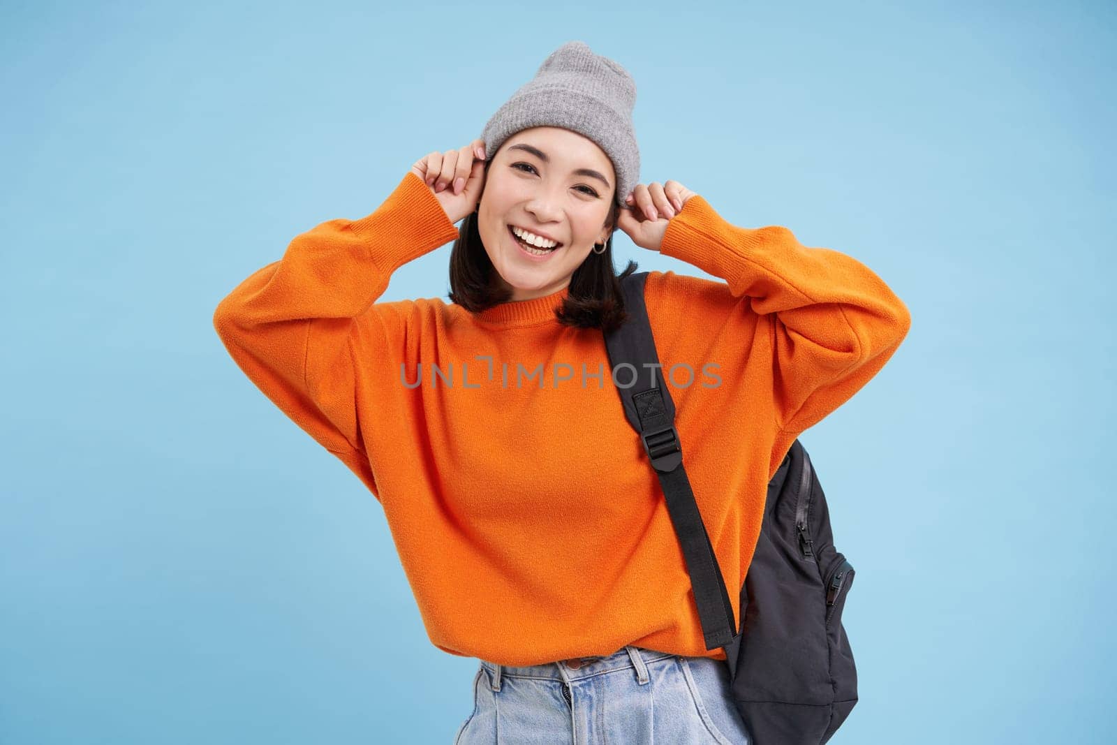 Stylish young asian woman in warm hat, walking with backpack, going somewhere with bag, standing over blue studio background by Benzoix