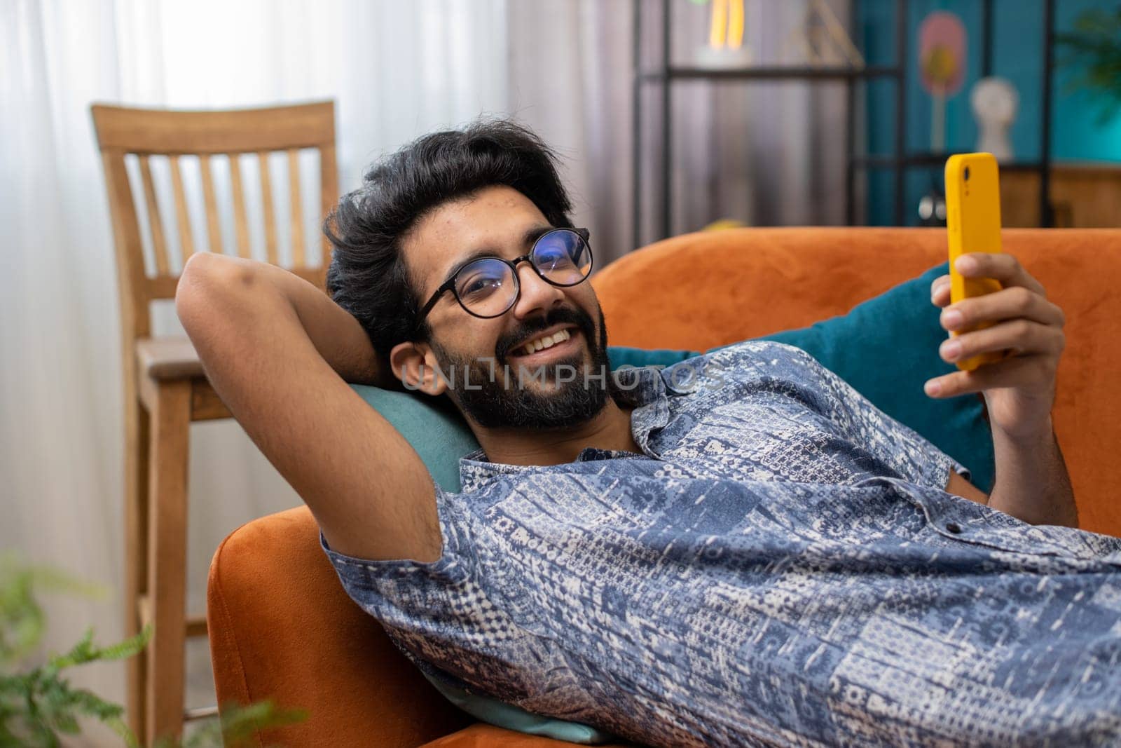 Indian man lying on couch uses mobile phone smiles at home living room apartment. Hindu young guy texting share messages content on smartphone social media applications online, watching relax movie
