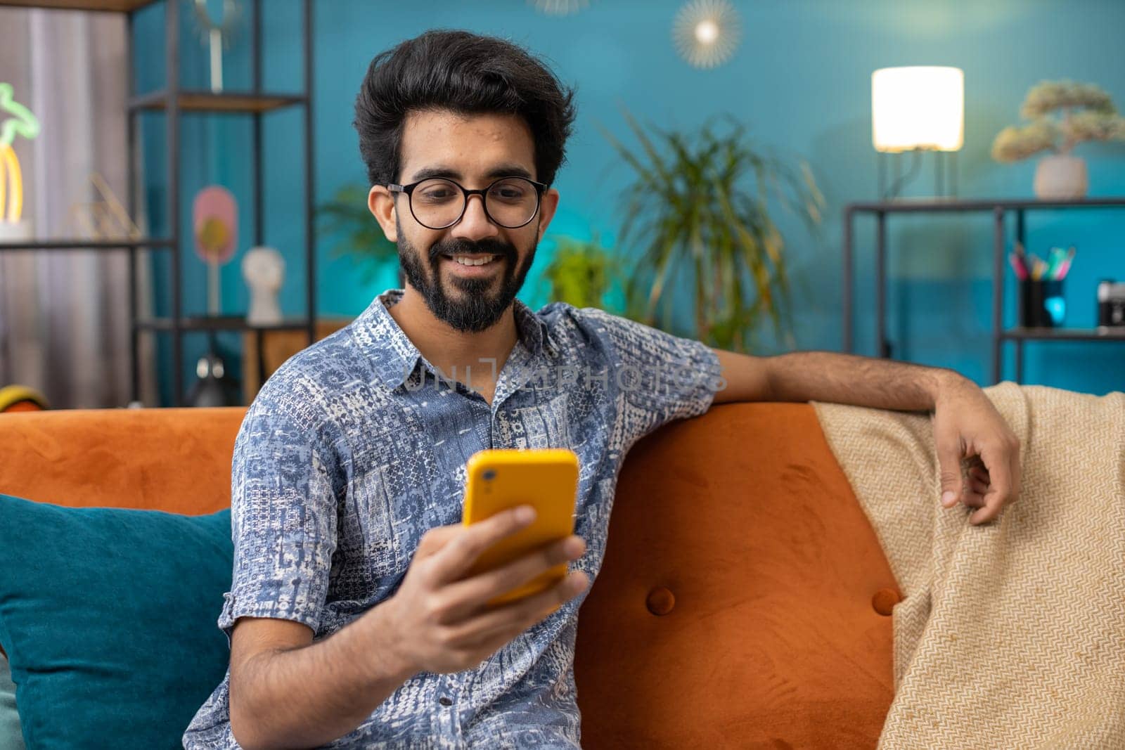 Indian man sitting on sofa uses mobile phone smiles at home living room apartment. Hindu young guy texting share messages content on smartphone social media applications online, watching relax movie