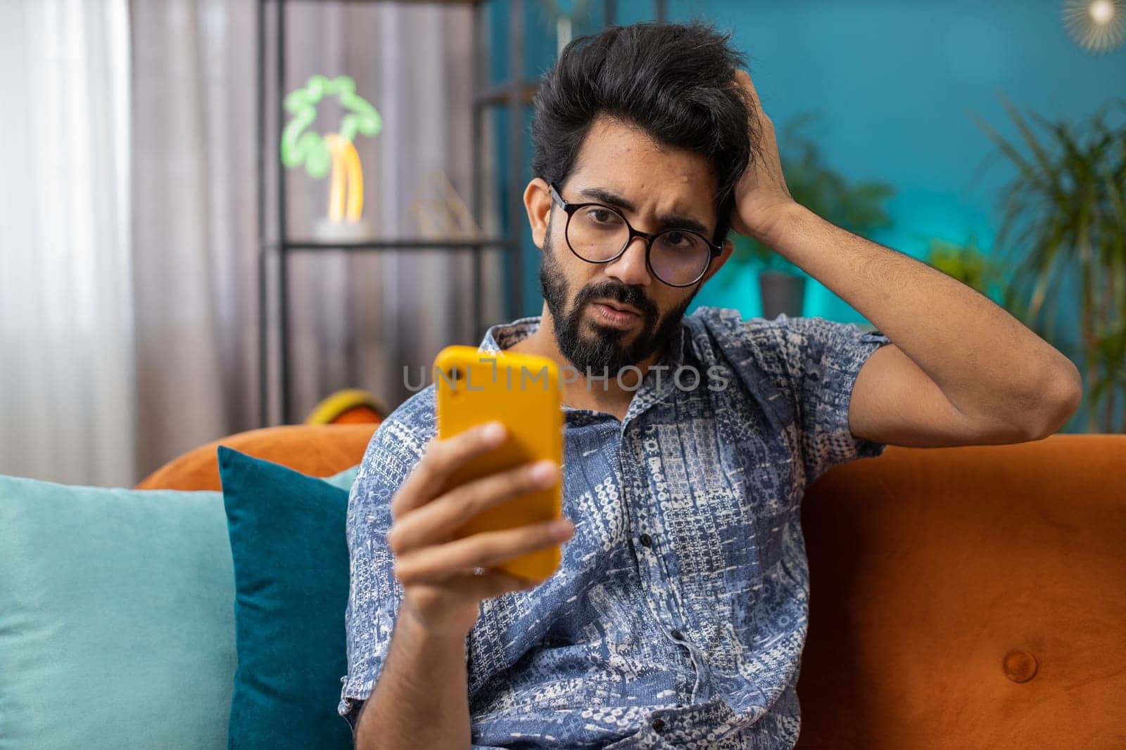 Sad displeased indian man use smartphone typing browsing, loses becoming surprised sudden lottery results, bad news, fortune loss, fail, deadline, virus. Young hindu guy at home living room on couch