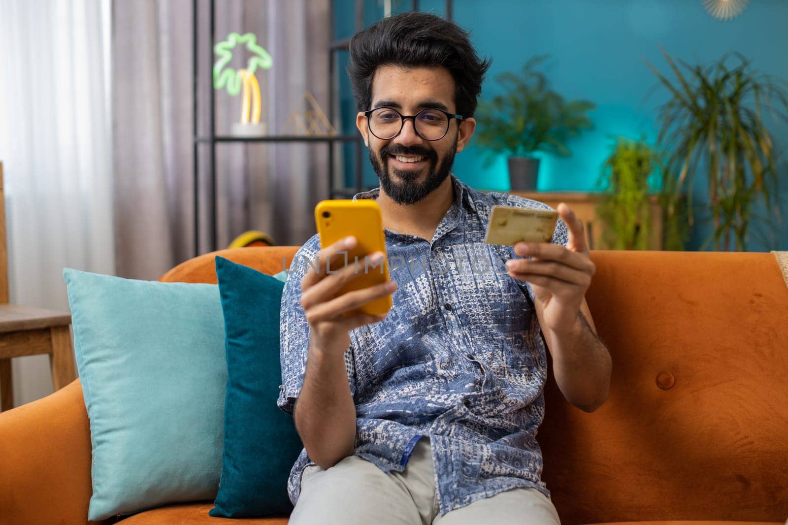 Indian man using credit bank card and smartphone while transferring money, purchases online shopping cashless, order food delivery at home apartment indoors. Happy hindu guy sitting in room on couch
