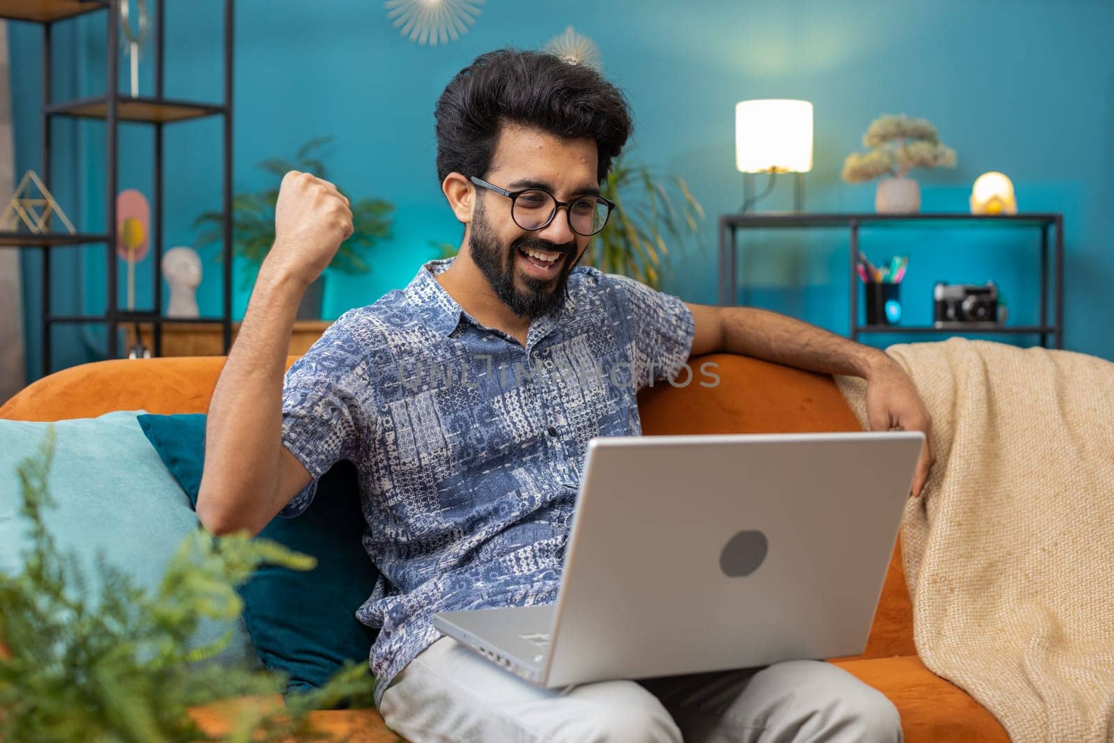 Surprised indian man guy using laptop computer, receive mail good news message, shocked by sudden victory celebrate lottery jackpot win purchases online shopping at home apartment on sofa. Lifestyles