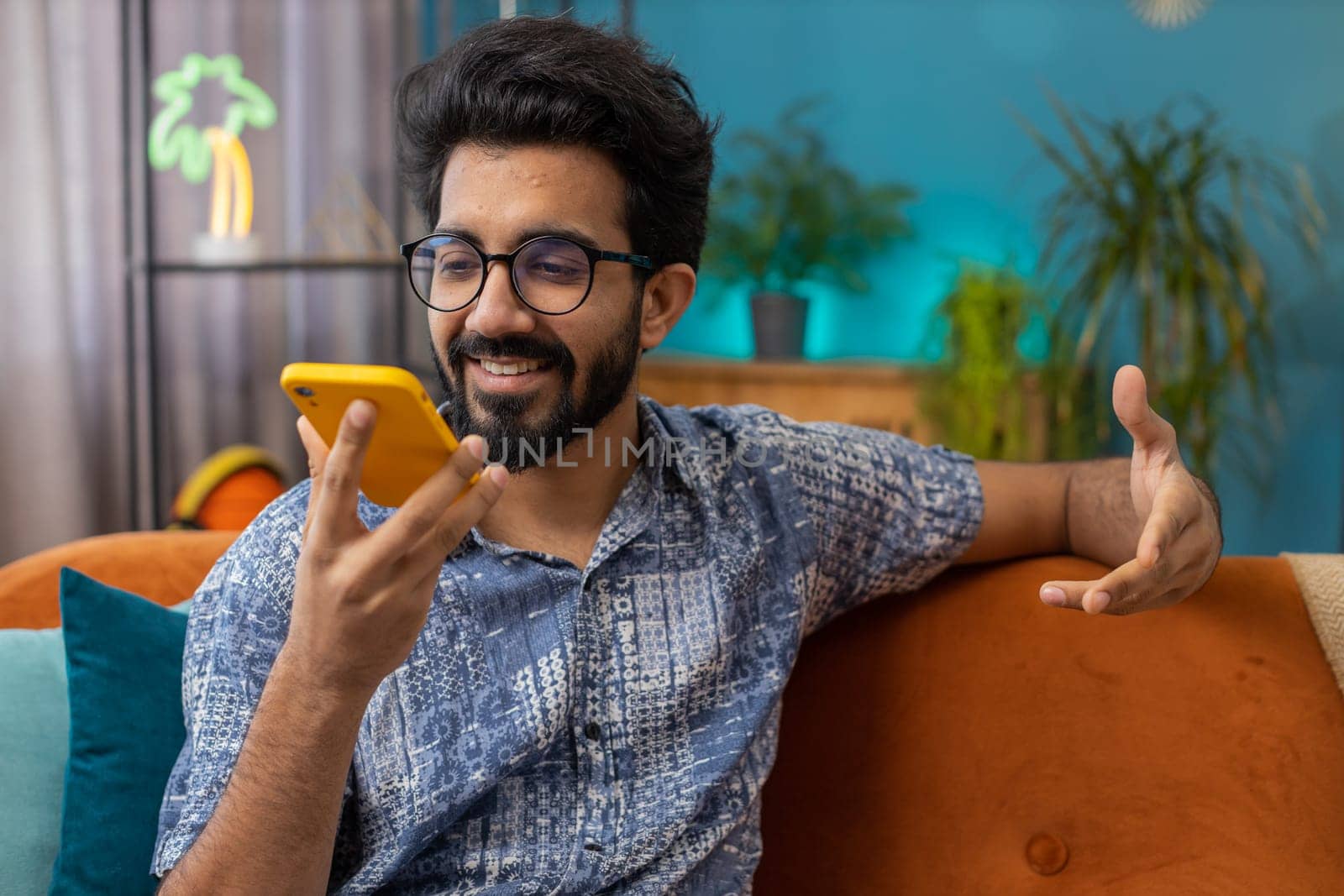Happy excited indian man making online phone call conversation with friends. Portrait of hindu guy enjoying mobile loudspeaker talking chatting communication at home apartment living room on couch