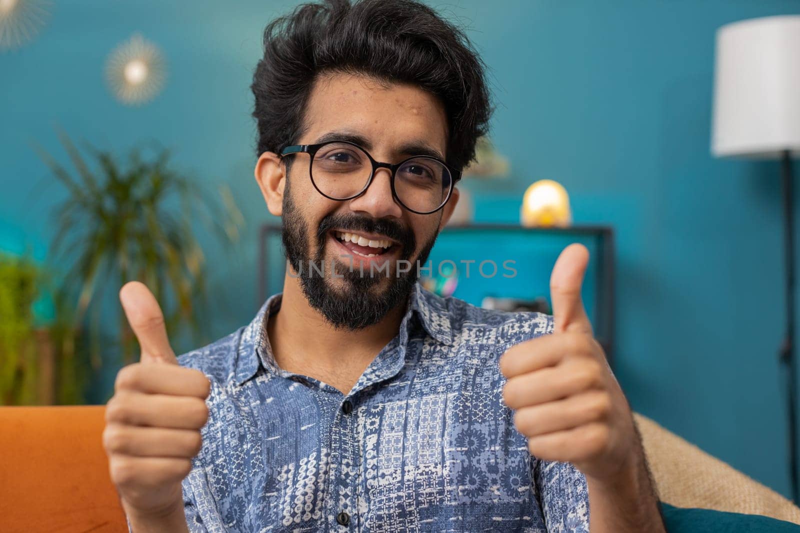 Like. Happy excited indian man looking approvingly at camera showing thumbs up, like sign positive something, good great news, positive feedback. Young hindu guy sitting on couch at home living room