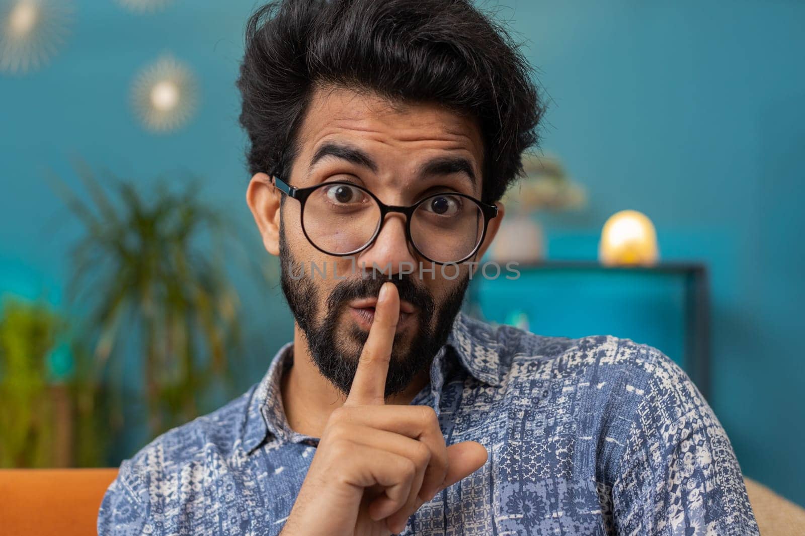 Shh be quiet, hush please. Indian man with presses index finger to lips makes silence hush gesture sign do not tells gossip secret. Young hindu guy at modern home living room sitting on couch. Gossip