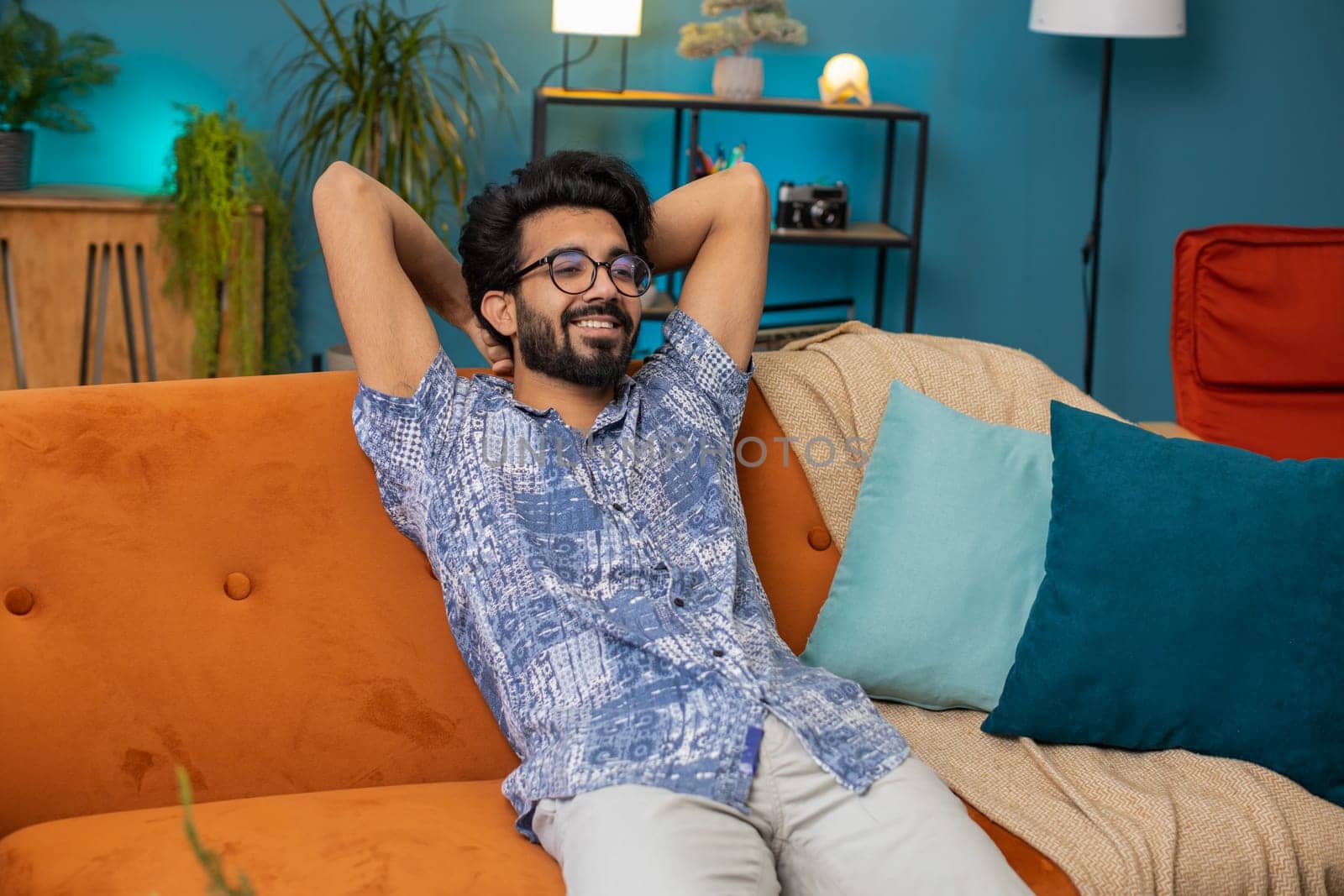 Tired indian man lying down on comfortable couch, taking a rest, daytime nap. Portrait of carefree hindu guy holding hands behind head relaxing, falling asleep at home apartment living room on sofa