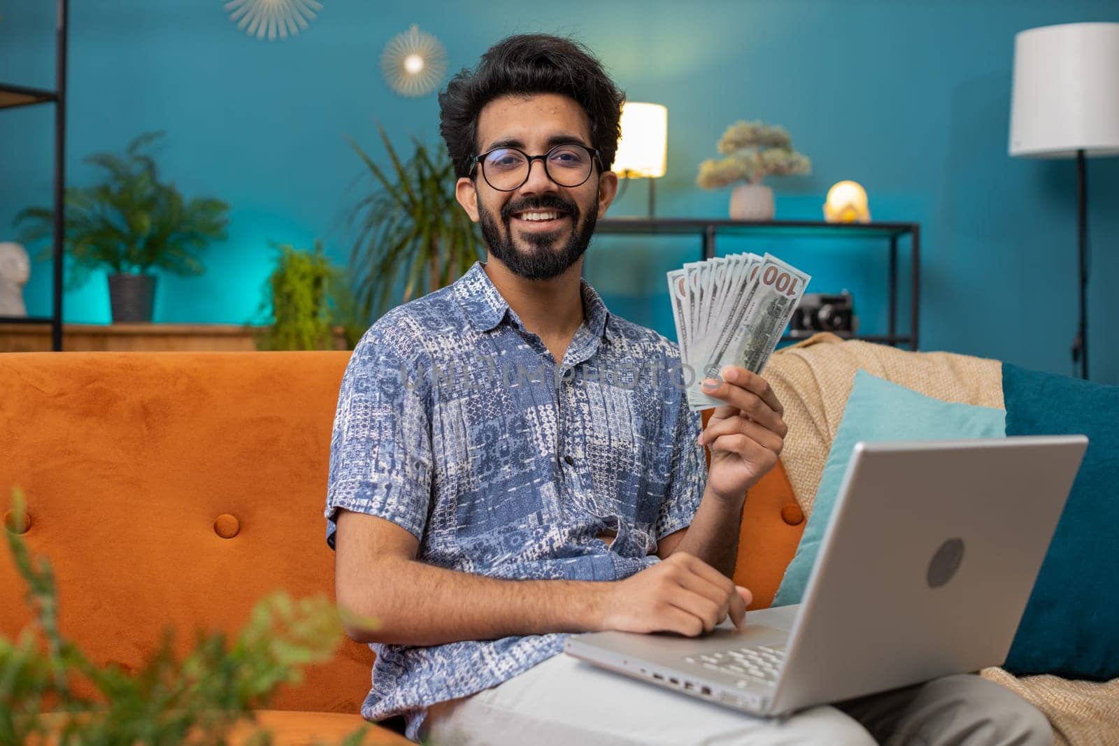 Planning budget. Rich happy indian man counting money cash use laptop computer calculate domestic bills at home room. Hindu guy satisfied of income earnings, saves money for planned vacation, gifts