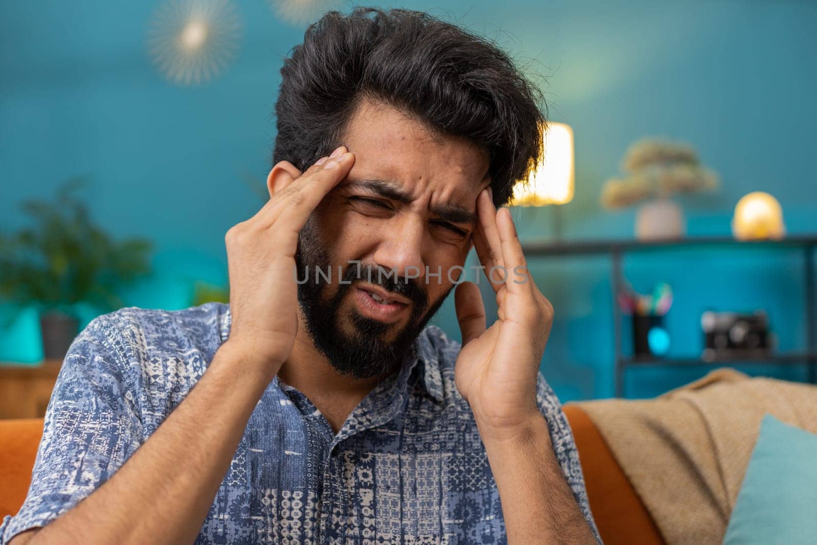 Displeased indian man rubbing temples to cure headache problem, suffering from tension and migraine, stress grimacing in pain high blood pressure at home room apartment. Hindu guy on couch. Lifestyles