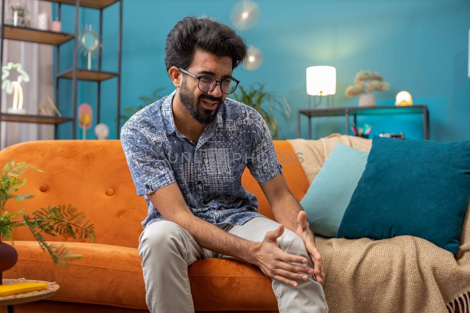 Young indian man suffer from painful severe knee joint ache, leg muscles, could not walk. Portrait of hindu guy massaging his knees illness cramps rheumatism at home room on sofa. Health care problem