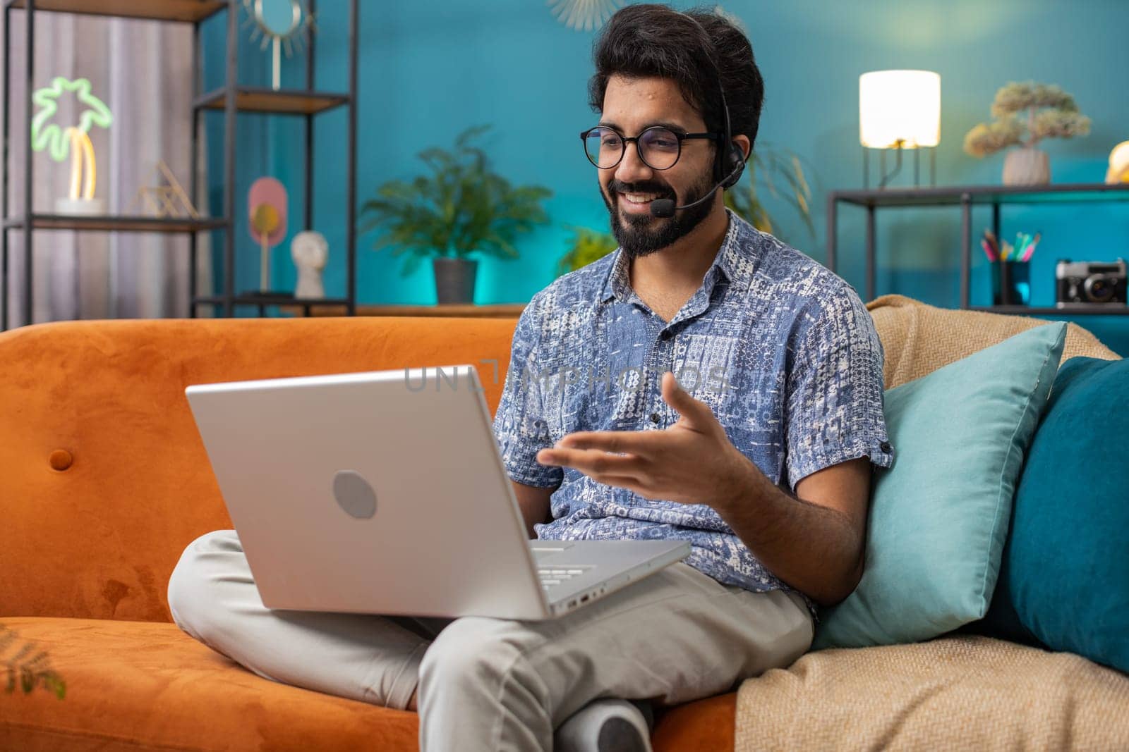 Indian man wearing headset, freelance worker, call center or support service operator helpline, having talk with client or colleague communication support waving hello. Hindu guy at home room on sofa