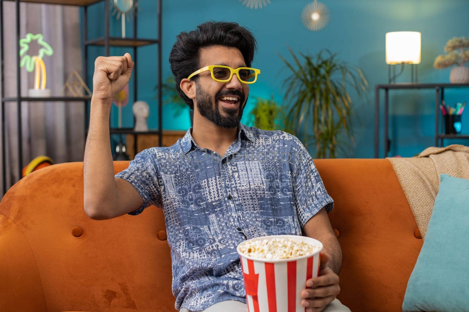 Excited indian man in 3D glasses sits on couch eating popcorn snacks and watching interesting TV serial sport game, film online social media movie content at home apartment. Guy enjoying entertainment