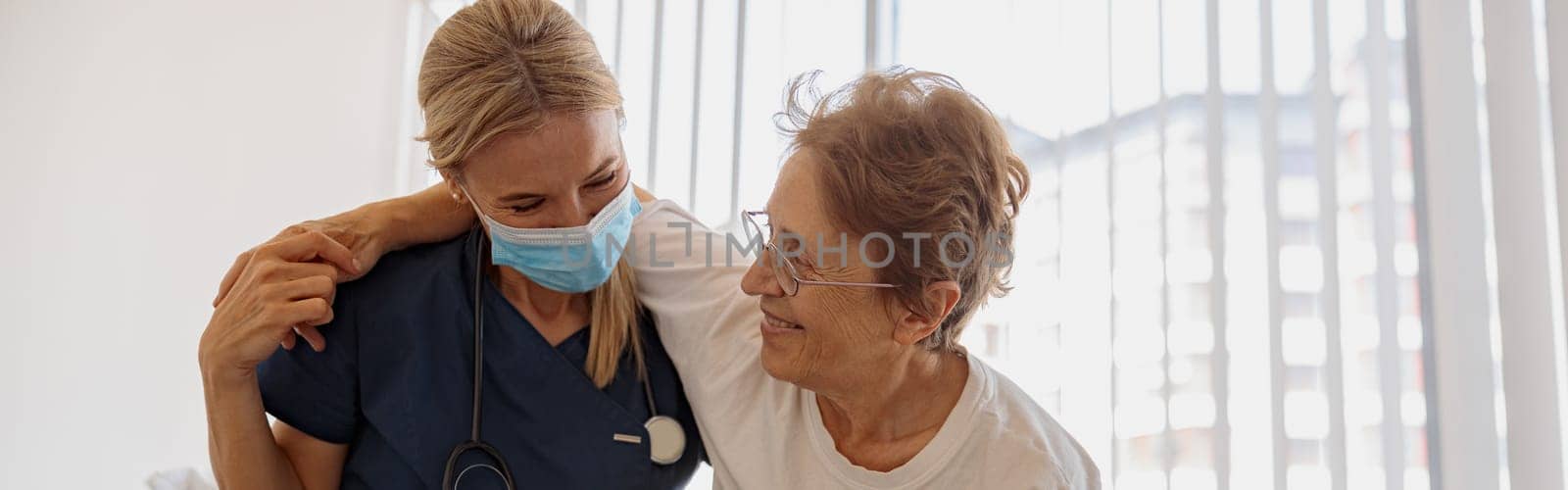 Female doctor in mask helping patient to get up from bed in the ward at hospital. High quality photo