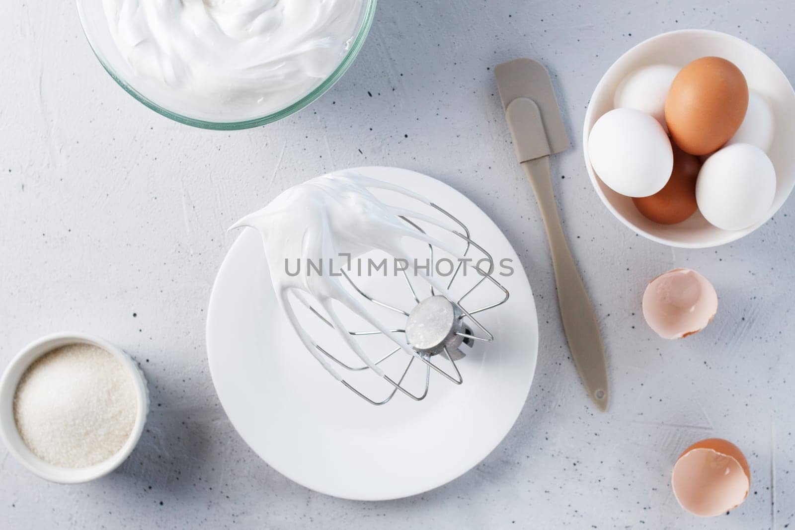 Whisked egg whites - whipped Italian meringue on a wire whisk and eggs on a gray background.
