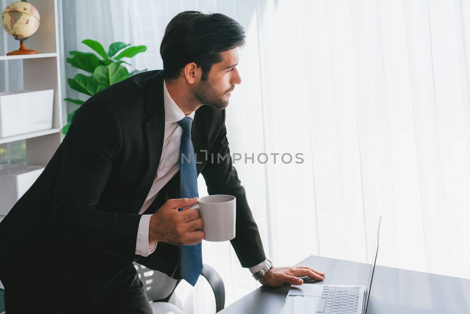 Modern professional businessman at modern office desk using laptop to work with coffee in his hand. Diligent office worker working on computer notebook in his office work space. fervent