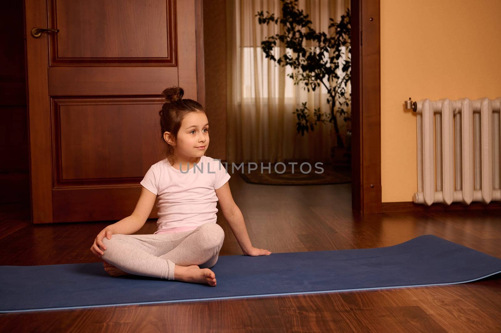 Adorable Caucasian 5 years old little child girl in active wear, sitting on yoga mat at cozy home interior. by artgf