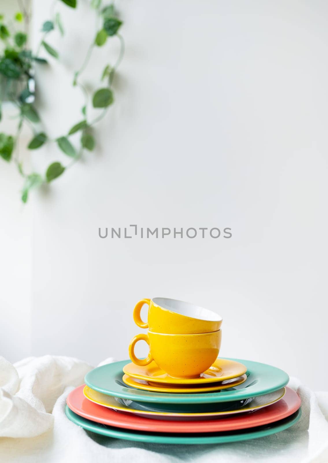 Empty yellow cups and brightly colored plates on a white table covered with a linen tablecloth, a flower pot on the wall in the background. Vertical photo. by sfinks