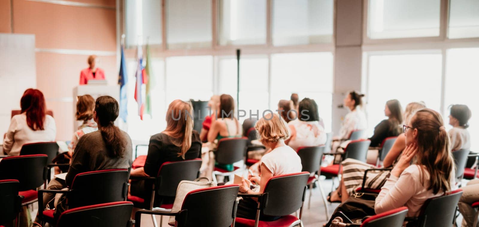 Business and entrepreneurship symposium. Female speaker giving a talk at business meeting. Audience in conference hall. Rear view of unrecognized participant in audience.