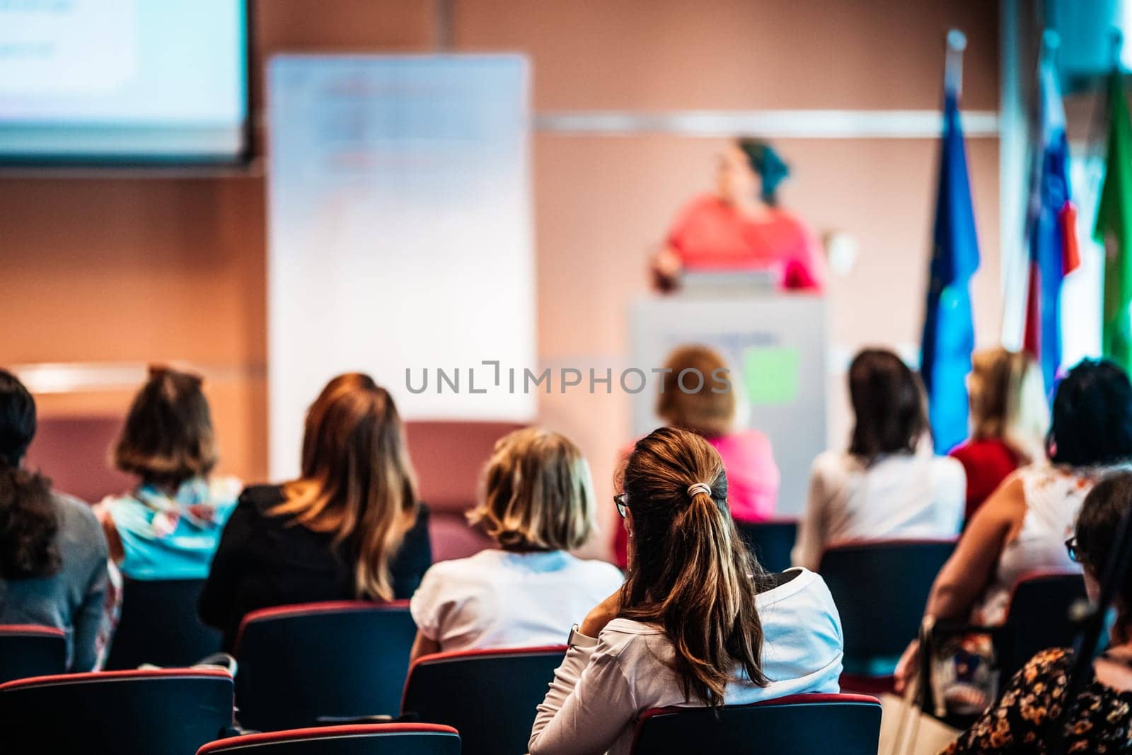 Business and entrepreneurship symposium. Female speaker giving a talk at business meeting. Audience in conference hall. Rear view of unrecognized participant in audience.