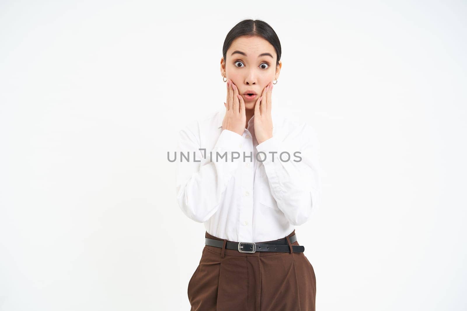 Corporate woman looks amazed, holds hands near face with surprised face expression, isolated on white background.