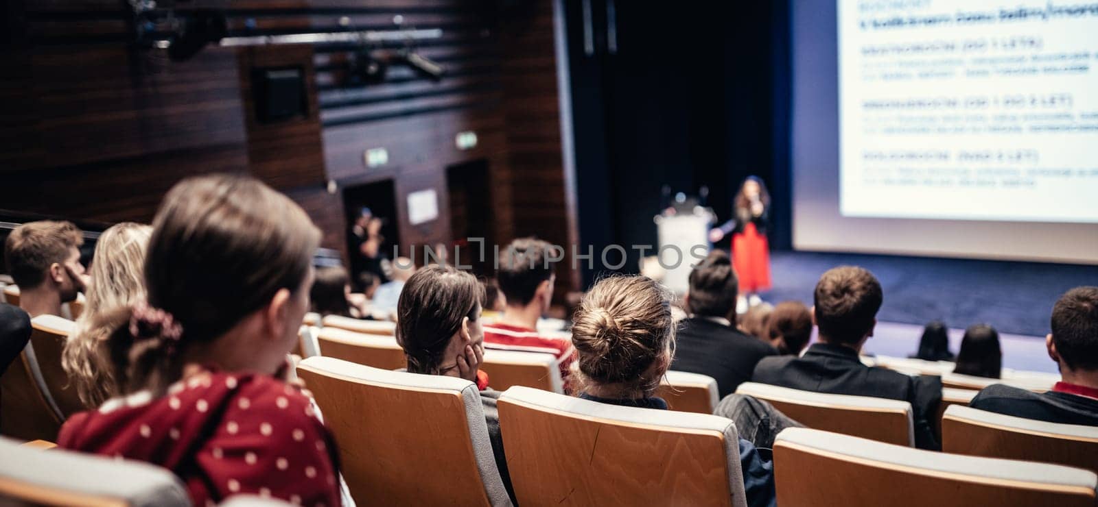 Business and entrepreneurship symposium. Female speaker giving a talk at business meeting. Audience in conference hall. Rear view of unrecognized participant in audience.