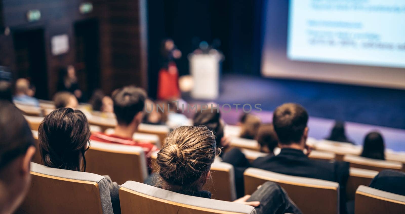 Business and entrepreneurship symposium. Female speaker giving a talk at business meeting. Audience in conference hall. Rear view of unrecognized participant in audience.