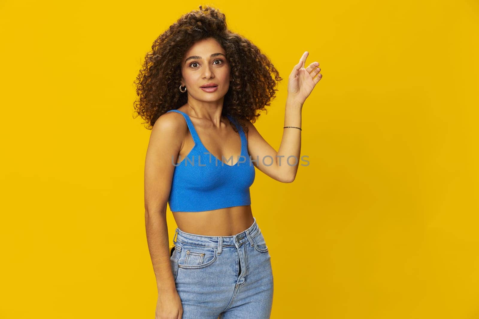 Woman with curly afro hair in a blue t-shirt on. yellow background signs with her hands, look into the camera, smile with teeth and happiness, copy space. High quality photo