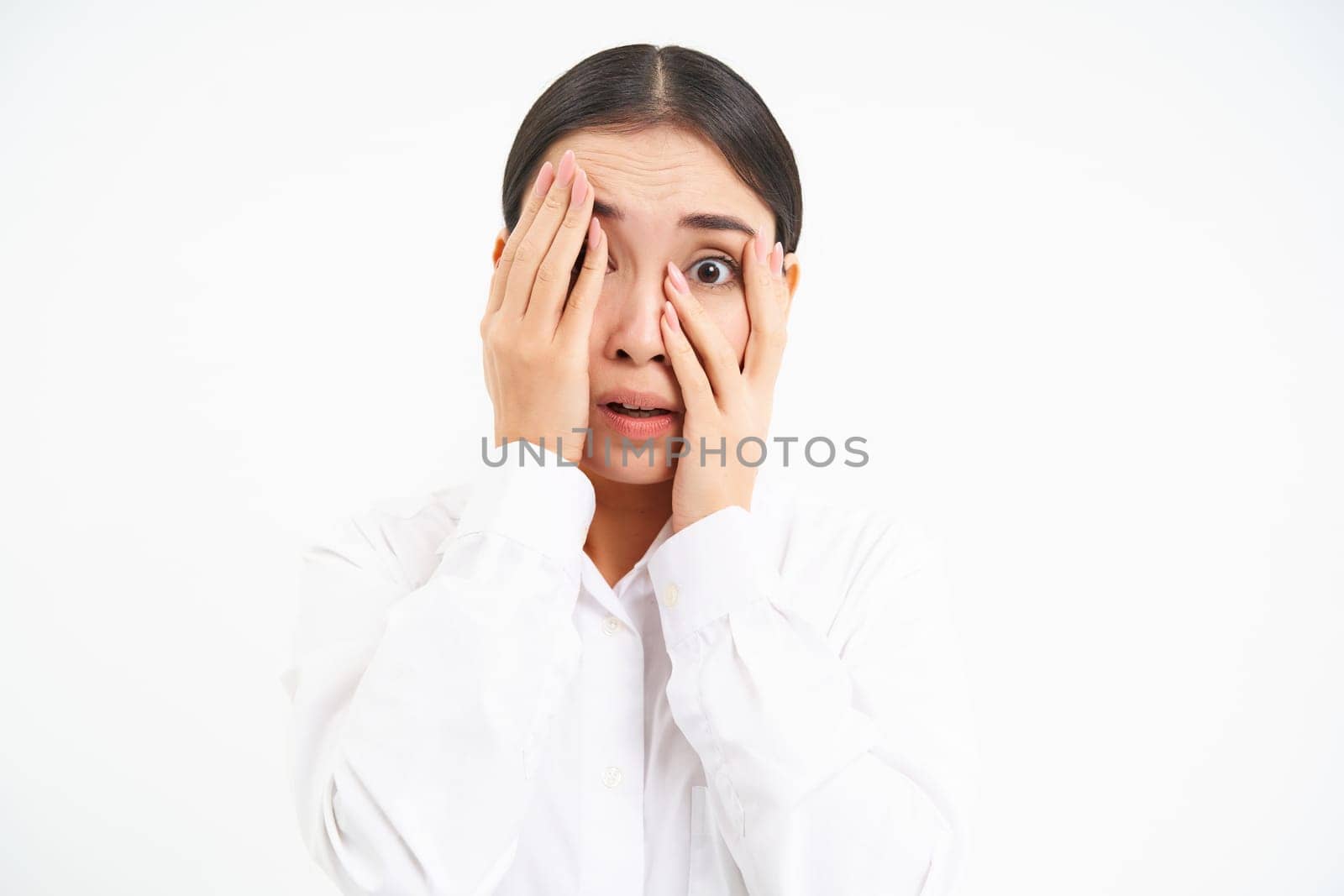 Japanese woman shuts face with hands, peeks through fingers with scared, terrified face, stands over white background.