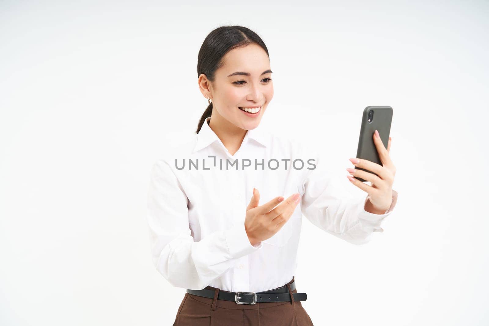 Portrait of corporate woman, manager video chats on mobile phone, looks at smartphone and leads a meeting online via smartphone app, white background.