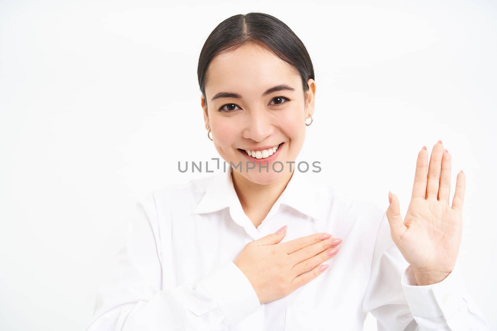 Friendly young businesswoman, asian lady raises one hand and puts palm on heart, makes promies, introduces herself, stands over white background.