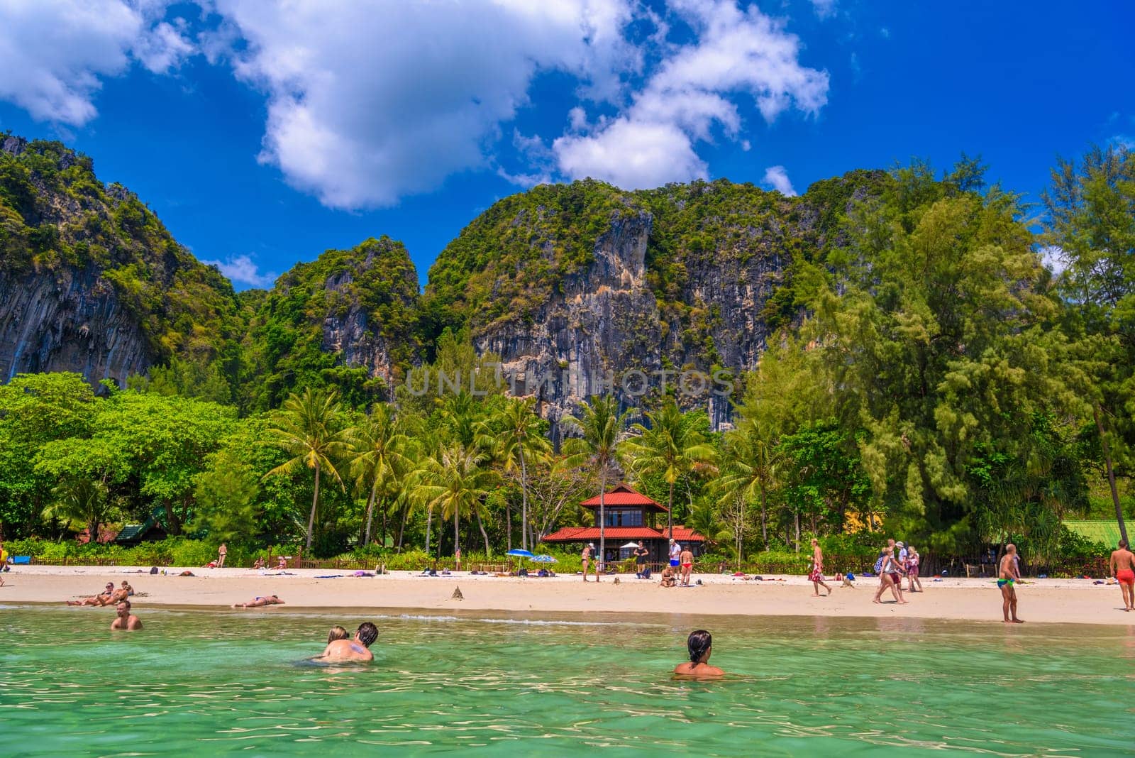 Bungalow house with red roof among coconut palms near the cliffs with people sunbathing and swimming in emerald water on Railay beach west, Ao Nang, Krabi, Thailand.