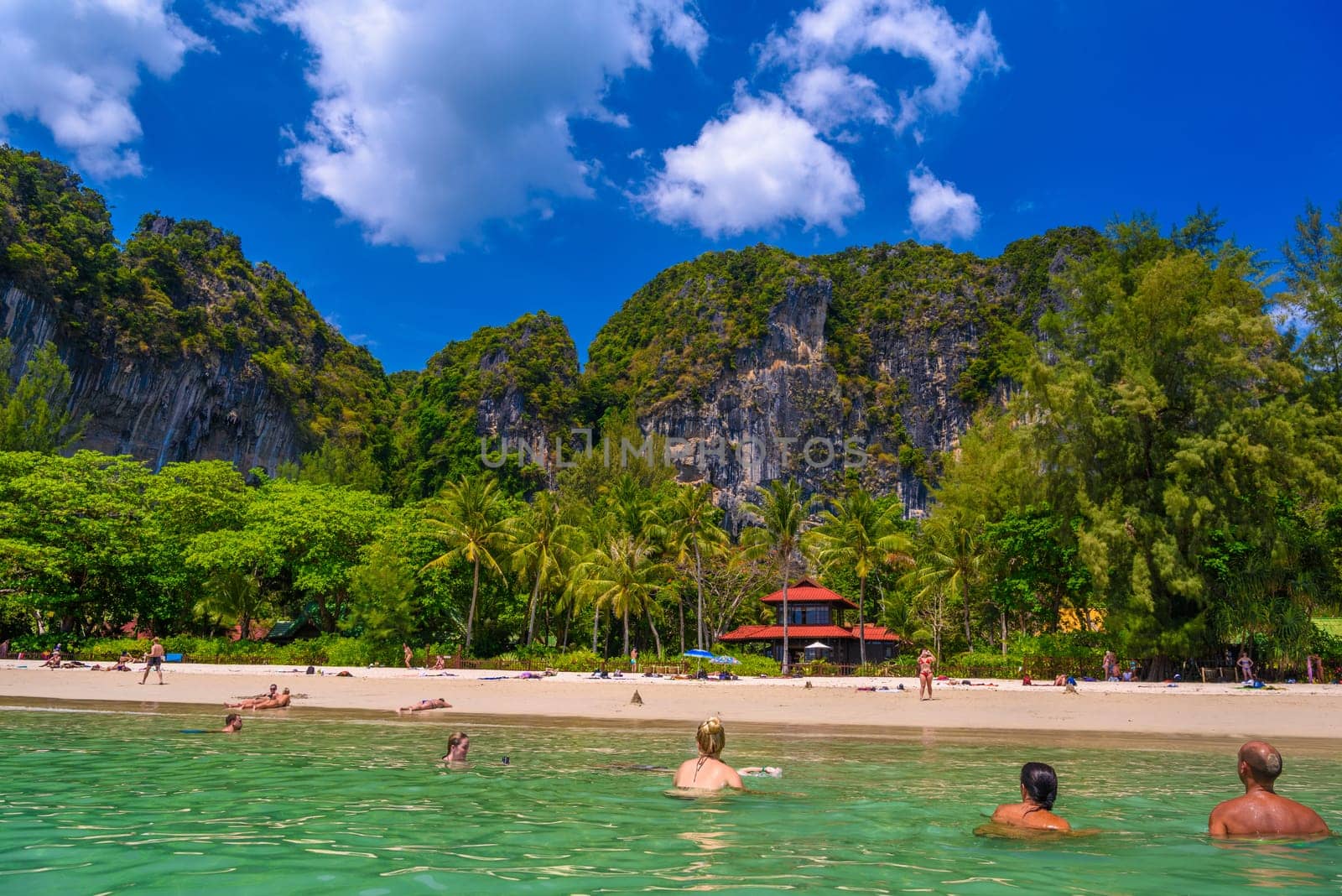 Bungalow house with red roof among coconut palms near the cliffs with people sunbathing and swimming in emerald water on Railay beach west, Ao Nang, Krabi, Thailand by Eagle2308