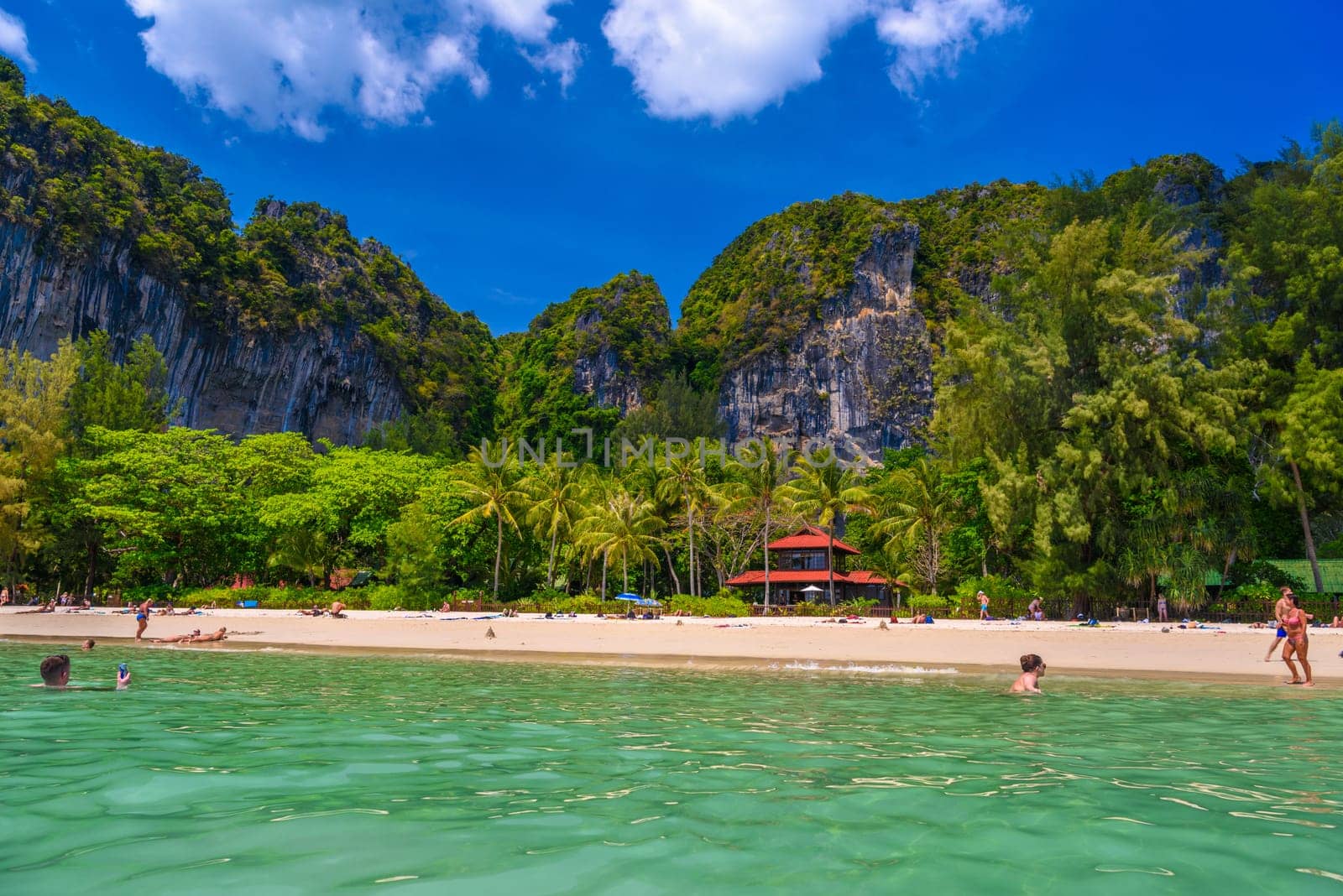 Bungalow house with red roof among coconut palms near the cliffs with people sunbathing and swimming in emerald water on Railay beach west, Ao Nang, Krabi, Thailand by Eagle2308