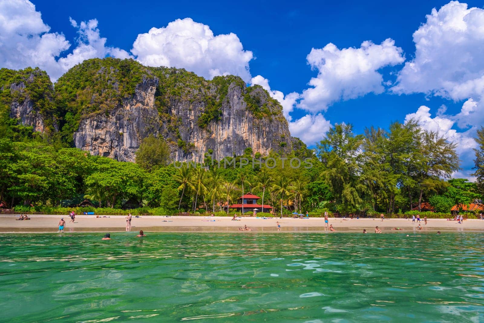 Bungalow house with red roof among coconut palms near the cliffs with people sunbathing and swimming in emerald water on Railay beach west, Ao Nang, Krabi, Thailand.